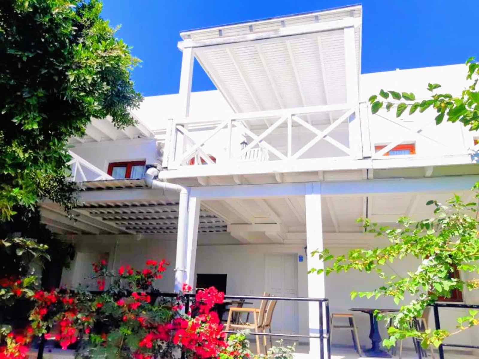 Beyaz iki katlı otel binası, ahşap pergolalı balkon ve kırmızı begonviller